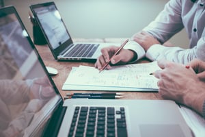 two people working at a table with laptops, paper and pens